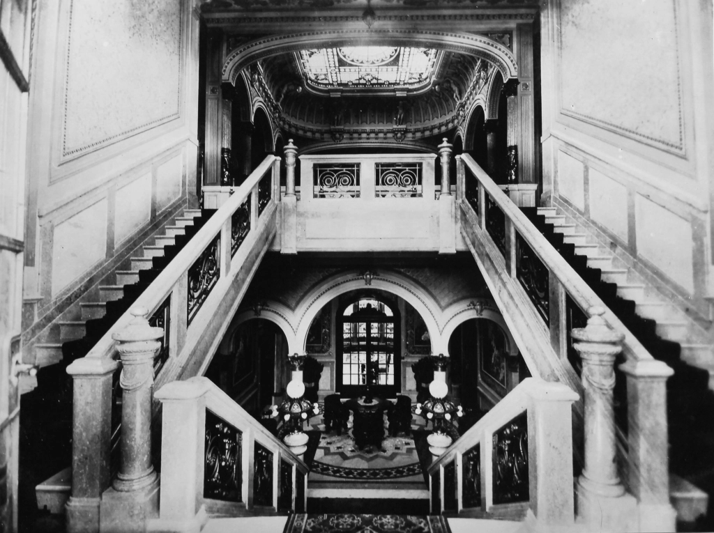 Fotografia interna do palacete, com duas escadas laterais que levam ao andar superior e uma escada central que leva ao andar inferior.