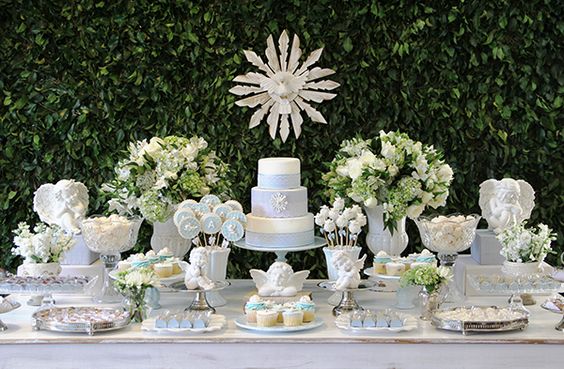 Mesa de bolo e doces do batizado, em branco e azul, além de ter muitas folhas e flores.