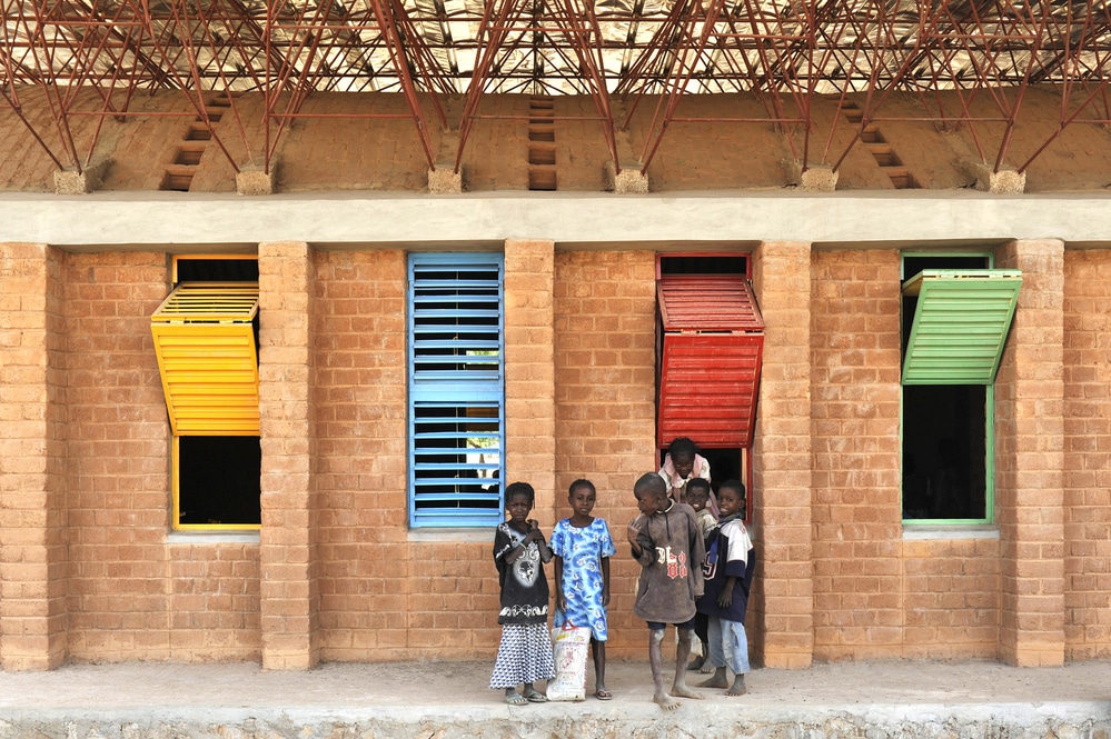 Imagem de uma das fachadas da Gando Primary School, também selecionada pela premiação, uma construção em tijolos aparentes e janelas coloridas. Há uma cobertura com estrutura de ferro que se apoia nos tijolos, permitindo entrada de ventilação e iluminação natural.