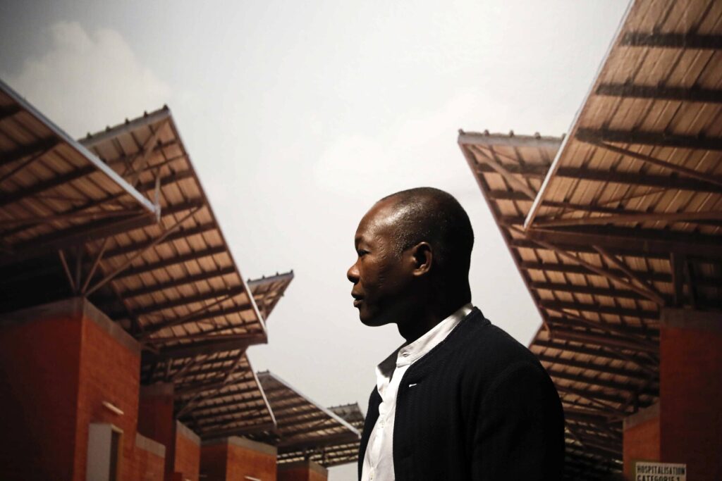 Fotografia de Francis Kéré observando uma de suas obras. Ao fundo há construções com tijolo aparente e telhados em direções variadas.