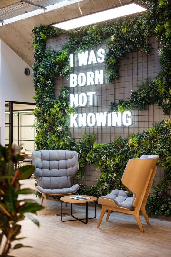 Ambiente comercial instagramável, com duas cadeiras acolchoadas na cor cinza, letreiro em led escrito "I WAS BORN NOT KNOWING" e jardim vertical.