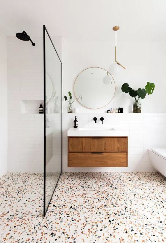 Banheiro moderno, majoritariamente branco com móvel de madeira e piso de granilite com fundo branco e mistura de cores com preto, cinza e marrom claro.
