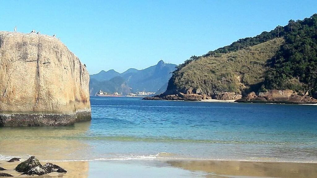 Piratininga, com mar azul, uma pedra grande bem em frente, na areia e os morros na parte de trás.