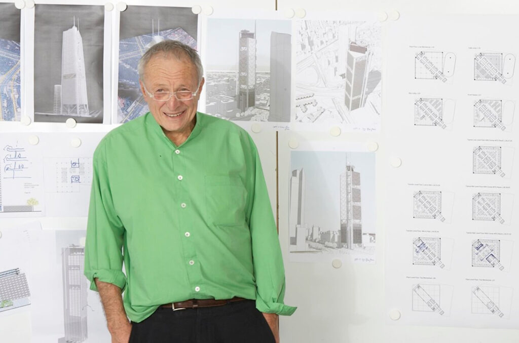 Imagem de Richard Rogers com camisa verde e calça preta, com algumas imagens impressas de projeto na parede que está atrás dele.