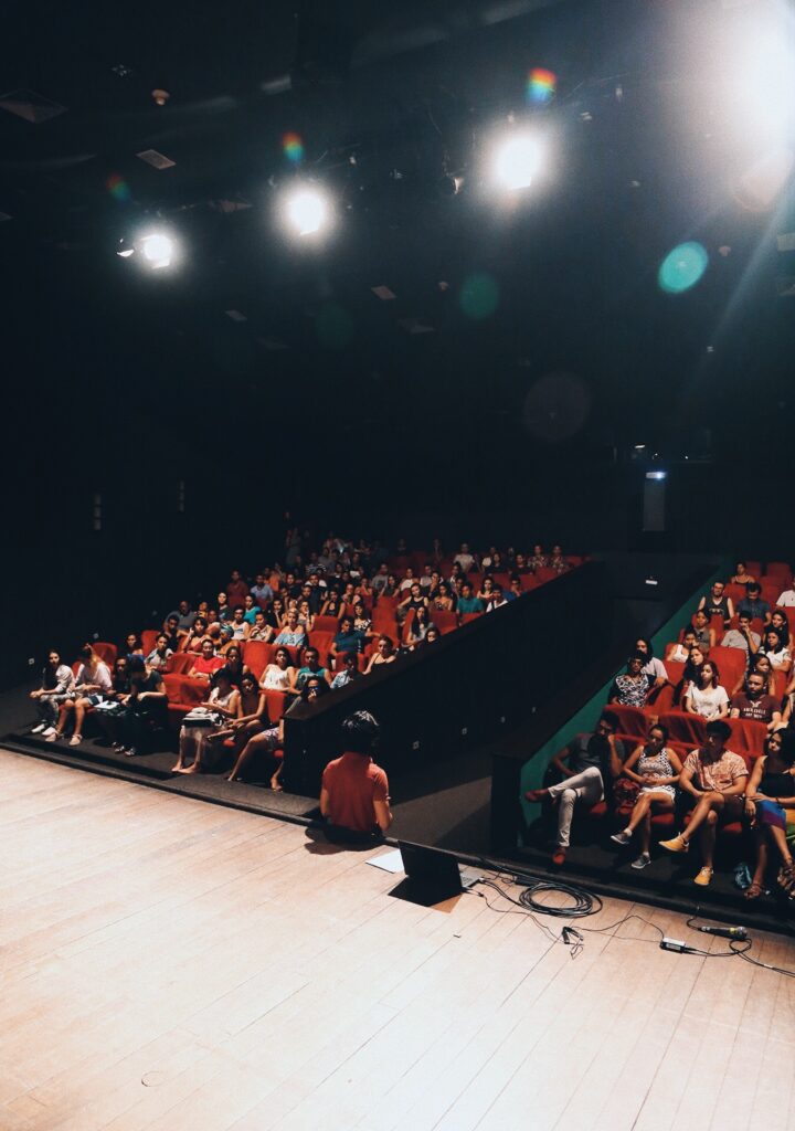 Imagem interna do teatro com pessoas sentadas nas cadeiras vermelhas vendo uma pessoa sentada no palco falando