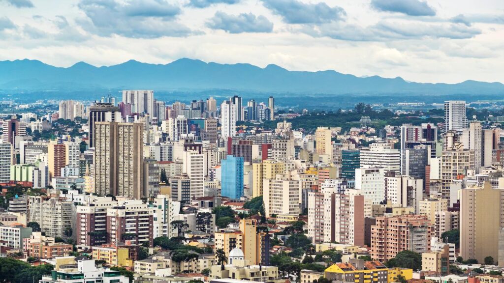 Cidade de Curitiba com prédios altos e ao fundo, montanhas e uma grande área verde.