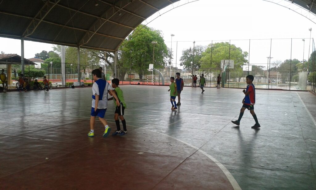 Quadra poliesportiva coberta com algumas crianças jogando futebol.