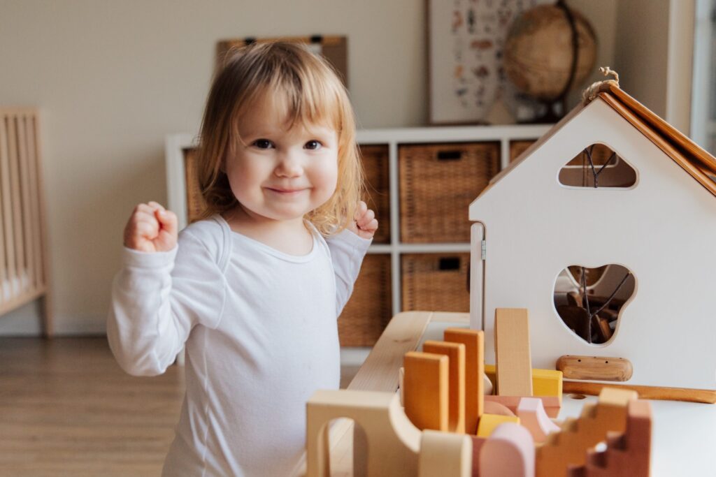 Criança com roupa branca ao lado de alguns brinquedos de madeira.