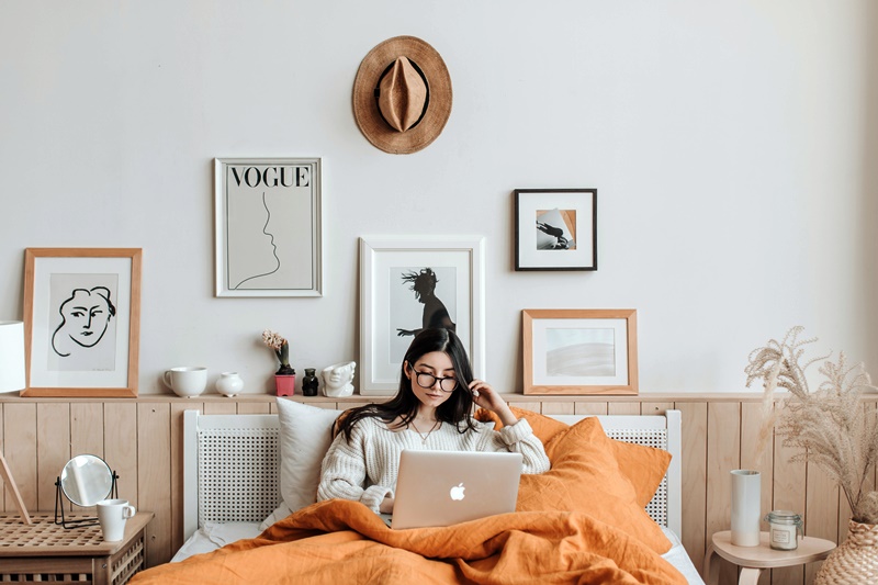Mulher com cabelo liso, escuro e comprido está sentada na sua cama usando seu notebook. Seu quarto é branco, possui alguns quadros artísticos na parede, em sua maioria em preto e branco, e as cobertas são laranja.