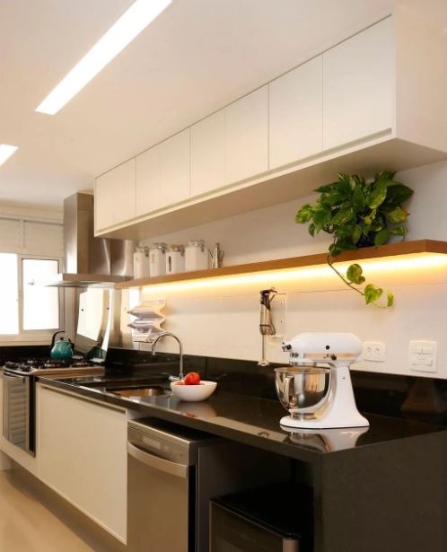 Aplicação da fita de LED sob a prateleira da cozinha, onde estão potes herméticos e uma planta para decoração. A prateleira está acima da bancada onde fica a pia e uma batedeira branca.