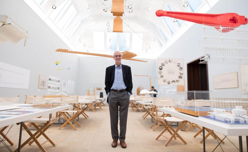 Fotografia de Renzo Piano andando por entre mesas nas quais estão dispostas diversas maquetes.