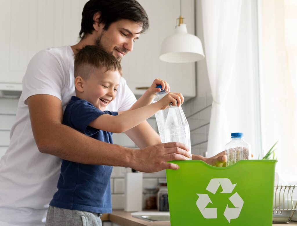 Homem de camiseta branca e menino pequeno de camisa azul colocando garrafas de plástico em uma caixa verde com o símbolo da reciclagem. 