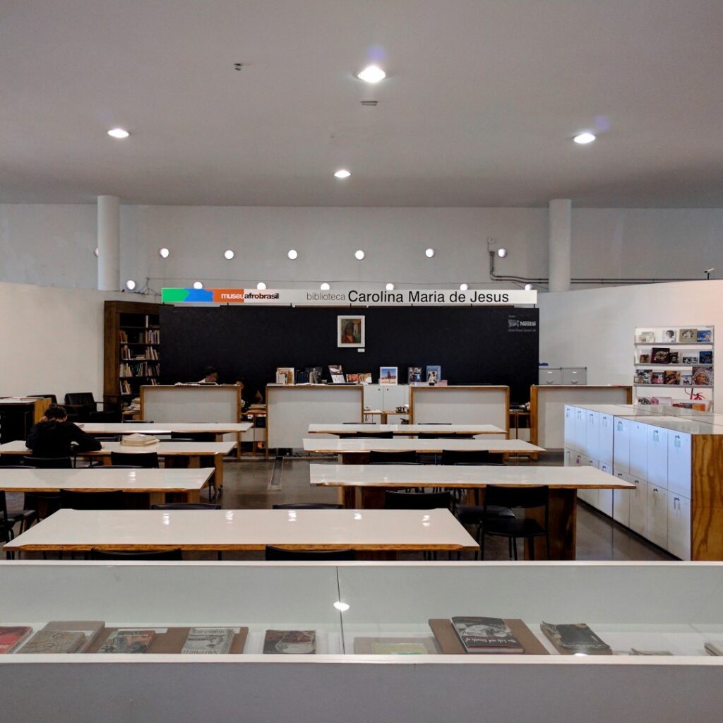 Fotografia da biblioteca, com amplas mesas para atividades e para leitura dispostas ao centro do ambiente.