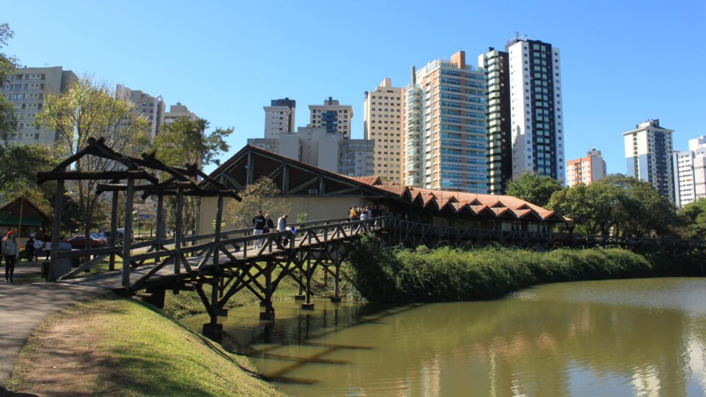 Imagem do Museu Botânico, que tem como acesso uma ponte de madeira, em frente ao museu, tem um lago e à trás estão os prédios da cidade. 
