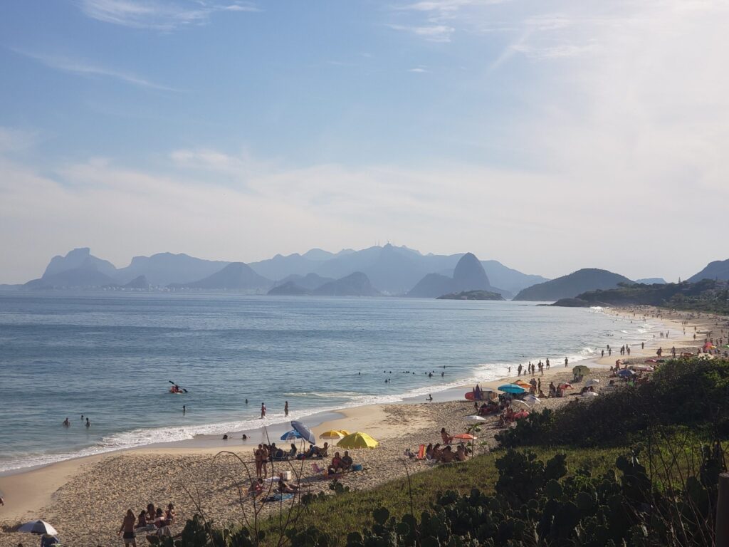 Camboinhas, com uma extensa faixa de areia e mar, com pessoas sentada na areia da praia tomando sol e outras aproveitando o mar. 