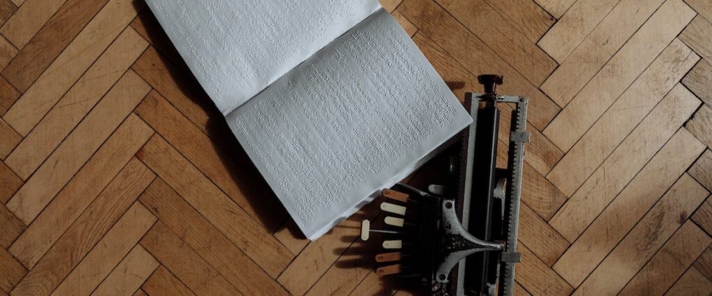 Piso de taco visto de perto com uma maquina de escrita em braile em cima e um caderno.